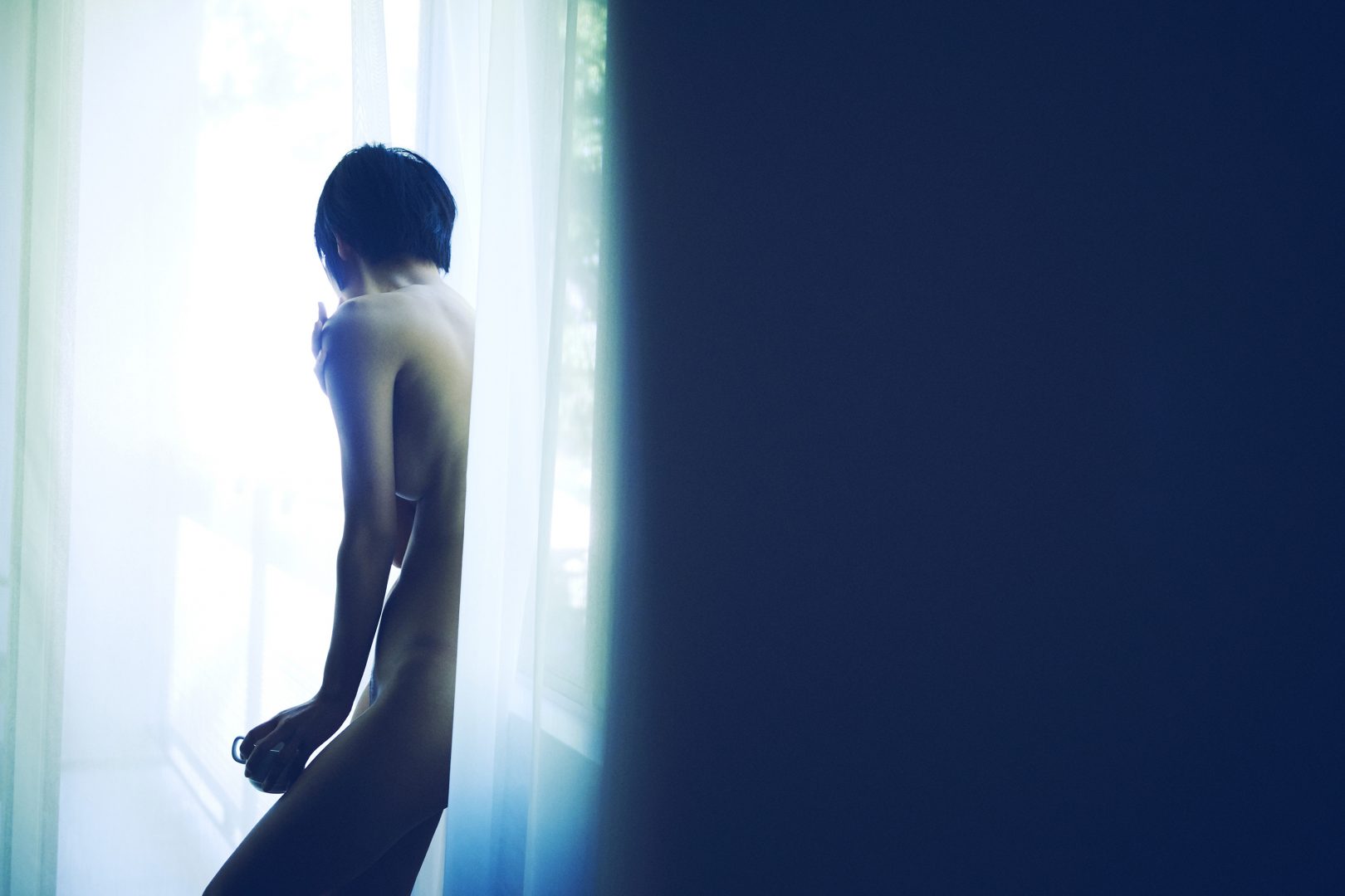 Naked girl with coffee cup in front of a window in hotel room by Stefan Rappo