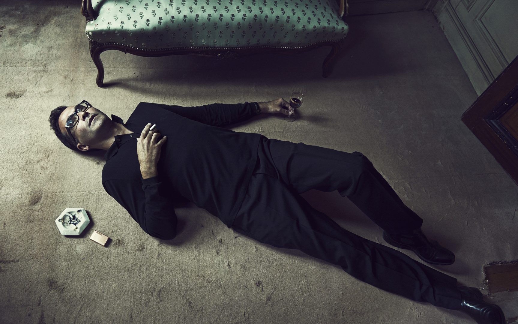 Man lying on floor with a cigarette and a glass of alcohol by Stefan Rappo